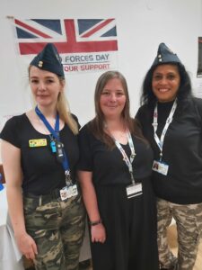Three women, two wearing RAF caps pose in front of an Armed Forces Day banner
