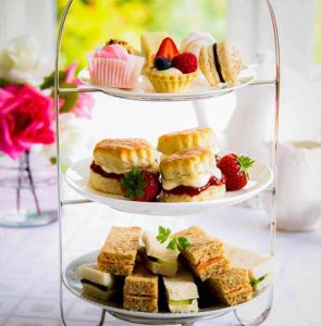  The image depicts a three-tiered afternoon tea stand. The top tier includes an assortment of delicate pastries, such as small fruit tarts and macarons. The middle tier features scones topped with clotted cream and strawberry jam, accompanied by fresh strawberries. The bottom tier presents an array of finger sandwiches with various fillings. The background shows a bright, elegantly set table with flowers, contributing to a classic afternoon tea setting.