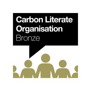 The image shows the logo of the "Carbon Literate Organisation Bronze" certification. The logo features a speech bubble with the text "Carbon Literate Organisation Bronze" inside it. Below the speech bubble are stylized figures holding hands, symbolizing community and collective action. The figures are depicted in a gold color, contrasting with the black speech bubble background and white text.