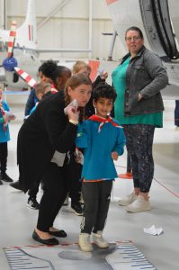 A Scout leader helps a young boy in blue Scouts uniform aim a paper plane down a large measuring tape placed on the floor in an aircraft hangar.