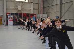 A group of children stand smiling in an aircraft hangar with their arms out in aeroplane poses.