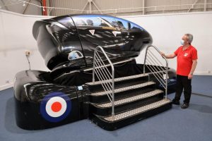 A staff member in a red shirt stands beside a black RAF-themed flight simulator pod with a staircase, as visitors inside watch a screen during the experience.