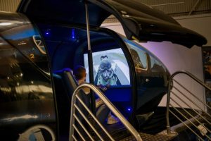 A child sits inside a dark, enclosed flight simulator pod watching a cockpit-view video of a jet in flight, with blue lighting and metal railings visible.