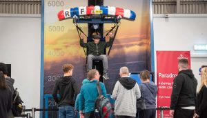 A young person wearing a VR headset experiences a paragliding simulator at the RAF Museum, suspended in a seated harness under a red, white, and blue canopy labeled “RAF”, while a crowd watches.