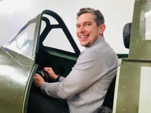 Smiling man seated in a Spitfire cockpit, holding the control stick and looking toward the camera.