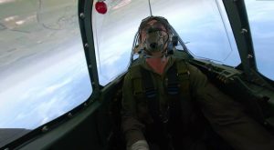 A pilot wearing a flight suit and oxygen mask performs an inverted maneuver inside a Spitfire aircraft, with the horizon flipped upside down through the cockpit canopy.