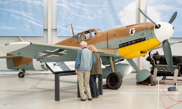 An elderly couple is observing a historical aircraft displayed in a museum. The aircraft is a World War II-era fighter plane with distinct markings, including a white nose, yellow underbelly, and camouflage paint. Behind the aircraft, there is an engine on display, and information plaques are placed in front of the plane for visitors to read.