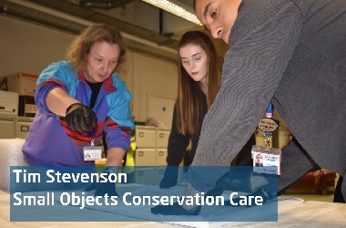 An image of a three people leaning over a working table with a paper artefact. They are mid conversation and one is making a gesture as if suggesting an action to take. A text box reads Tim Stevenson, Small Objects Conservation Care
