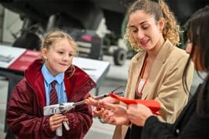 A teacher wearing a lanyard demonstrates a hands-on STEM activity to a young girl in school uniform.