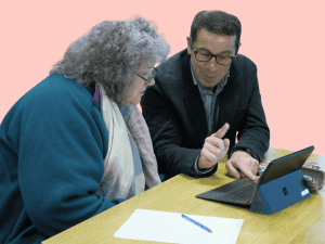 A lady and man sit at a desk looking at a tablet device. The man is explaining the device to the lady