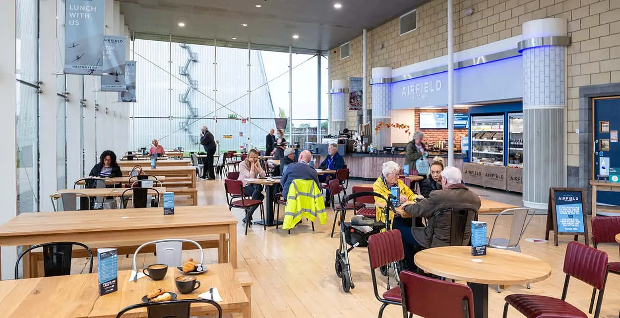 People sit at tables inside the Airfield Kitchen at the RAF Museum Midlands. Some are eating and talking, while others walk by the counter. The café has large windows, light wood floors, and a casual atmosphere.