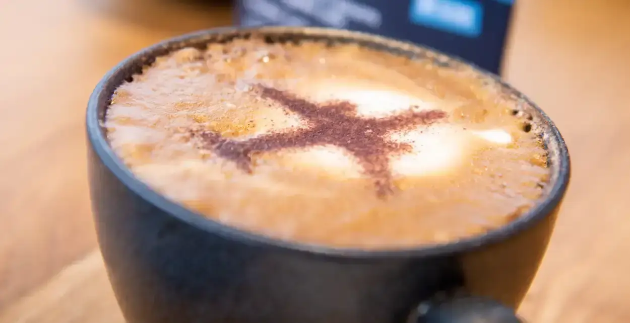A cup of cappuccino on a wooden table with an aeroplane shape dusted in cocoa powder on the frothy milk surface.