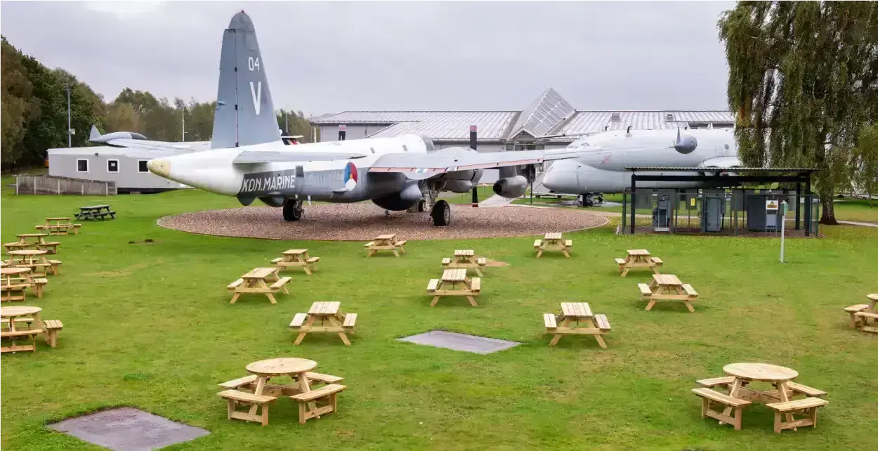 Several wooden picnic tables are arranged on a grassy area in front of a large military aircraft on display at the RAF Museum Midlands.