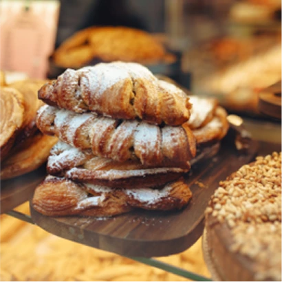 A stack of flaky croissants dusted with powdered sugar sits on a wooden board in a bakery display case, surrounded by other pastries.