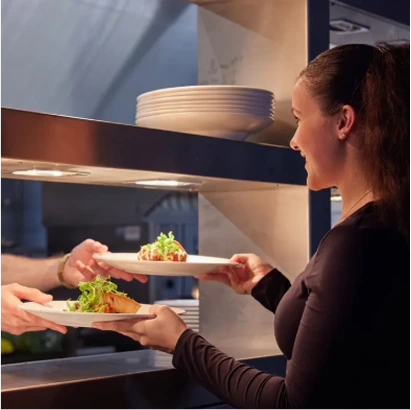A woman receives two plates of food from a kitchen counter, with stacked white plates visible above the serving area.