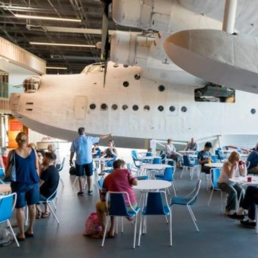 People gather and interact beneath a large historic seaplane display in a spacious indoor setting at the RAF Museum London.