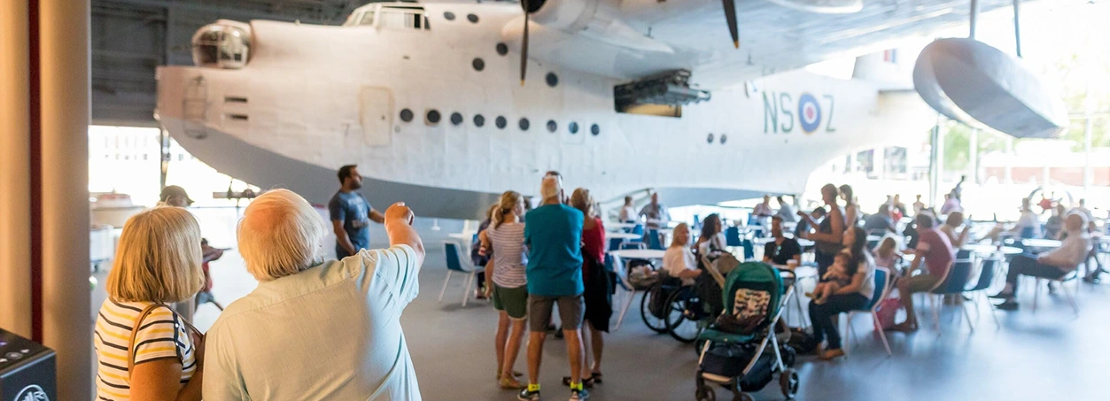 People gather and interact beneath a large historic seaplane display in a spacious indoor setting at the RAF Museum London.