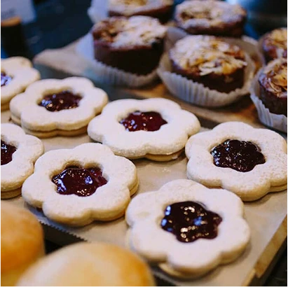 A close-up of flower-shaped cookies with powdered sugar and red jam centres, arranged on a tray. Muffins with almond slices are visible in the background.