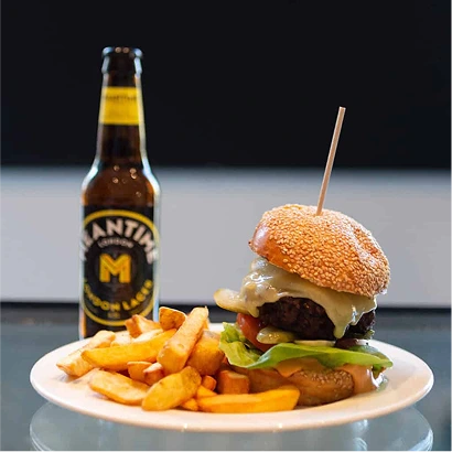 A plate with a cheeseburger topped with lettuce, tomato, and a sesame seed bun, served with thick-cut fries. A bottle of Mantins Major Lager beer is in the background.