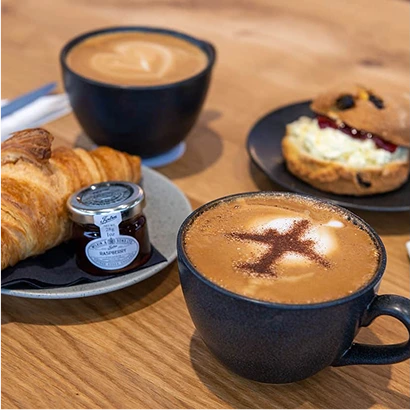 A wooden table with two cups of coffee, a croissant with raspberry jam, a scone filled with cream and jam, and cutlery in the background. One coffee has a chocolate aeroplane design on the froth.